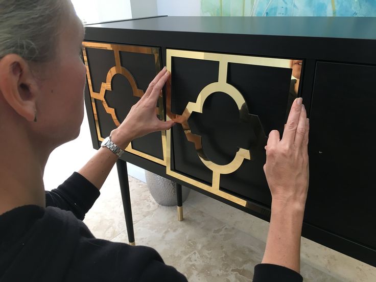 a woman is working on a cabinet with gold and black paint, while another person holds their hand up to the door