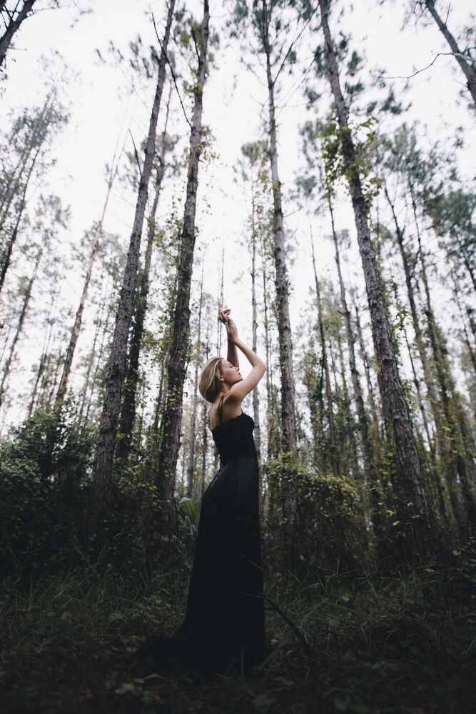 a woman standing in the middle of a forest with her arms raised up to the sky