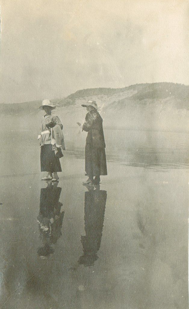 two people are standing in the water with their reflection on the wet ground and one person is wearing a hat