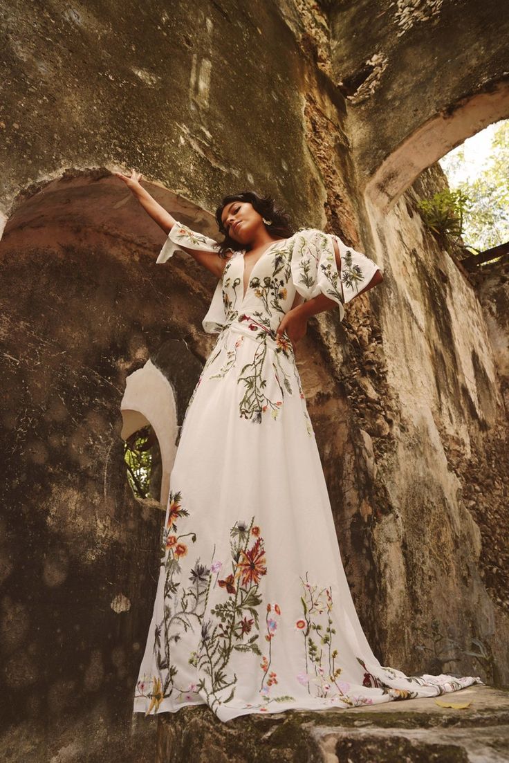 a woman in a long white dress leaning against a stone wall with her hands on her hips