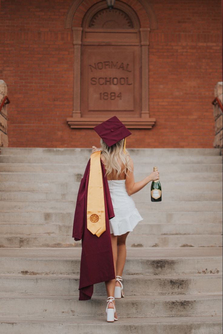 a woman in a graduation gown is holding a bottle of wine and standing on some steps