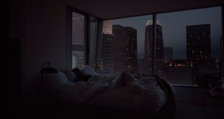 an unmade bed in front of a large window overlooking the city skyline at night