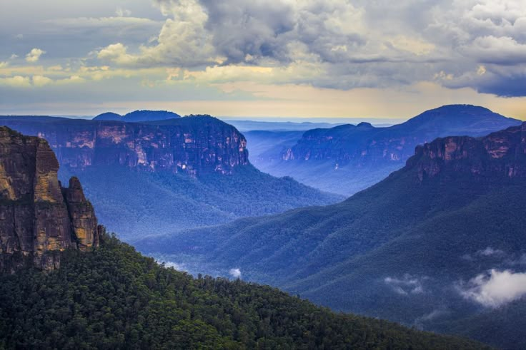 the blue mountains are surrounded by lush green trees and cloud filled sky with clouds in the distance