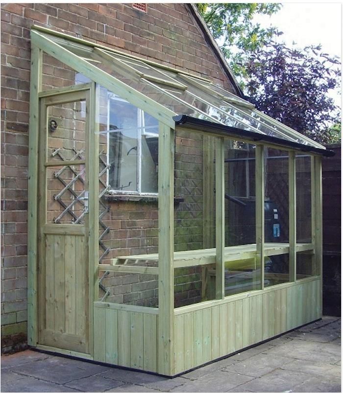 a wooden greenhouse with glass doors and windows on the side of a brick building next to a tree