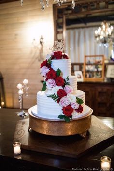 a wedding cake with red and white flowers on it sits on a table in front of candles