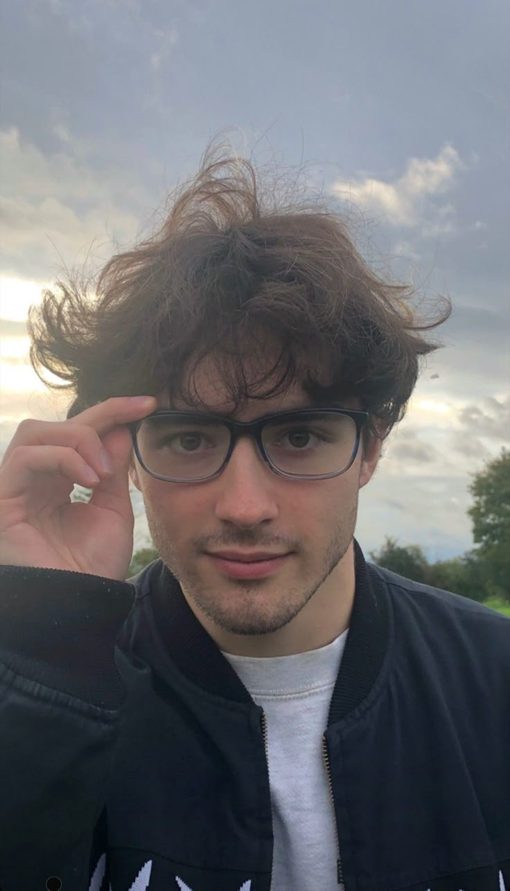 a man wearing glasses and a jacket with his hair blowing in the wind while looking at the camera