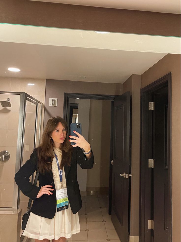 a woman taking a selfie in front of a bathroom mirror while wearing a white dress and black blazer