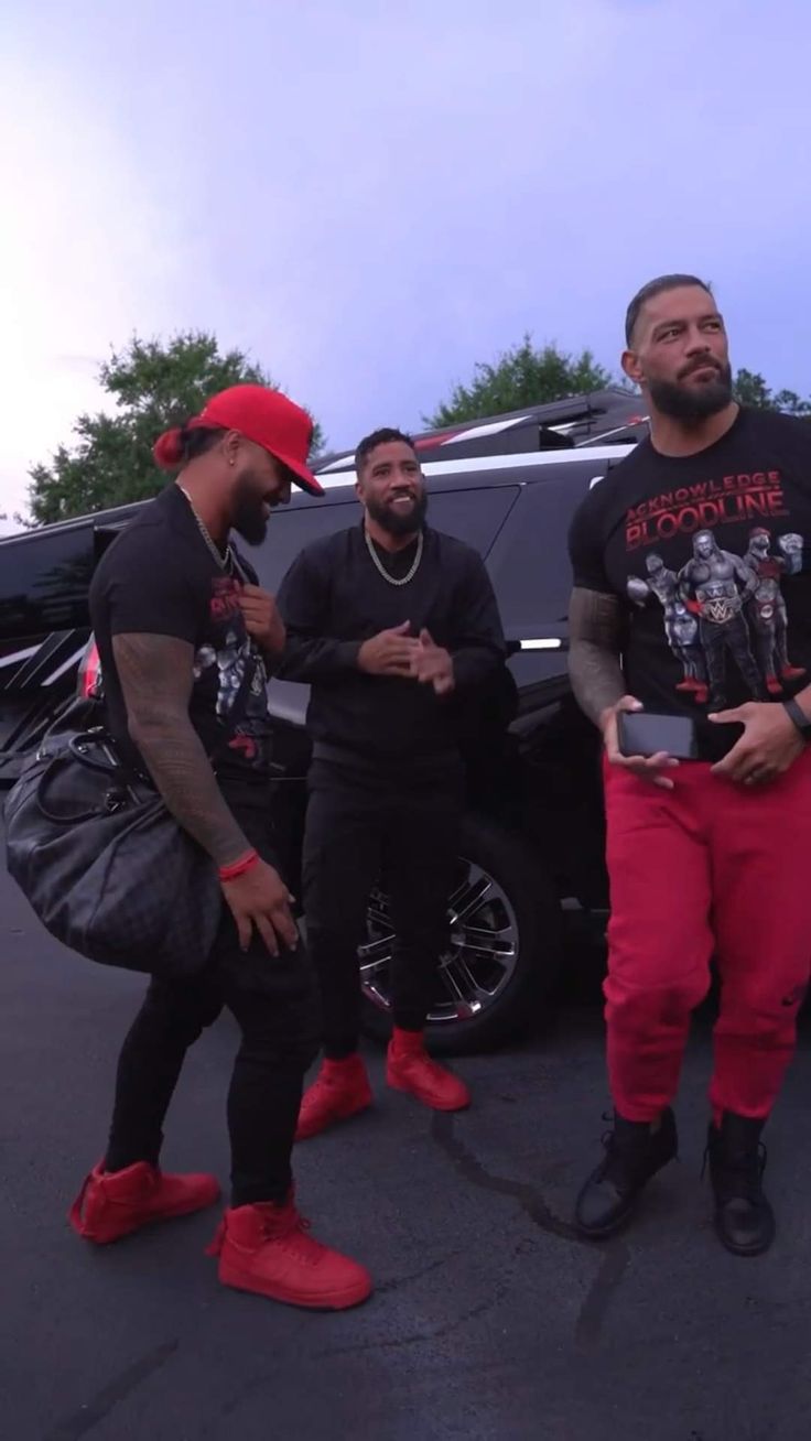 three men standing in front of a black car wearing red pants and matching t - shirts