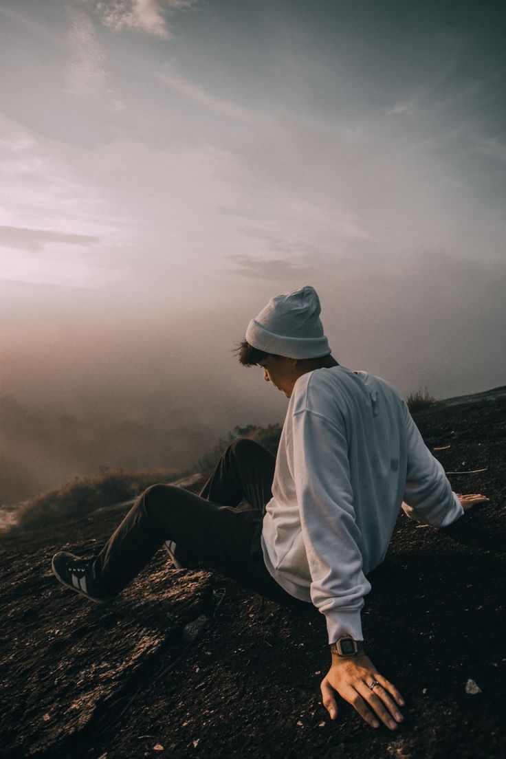 a man sitting on top of a mountain with his hands in the air while wearing a hat