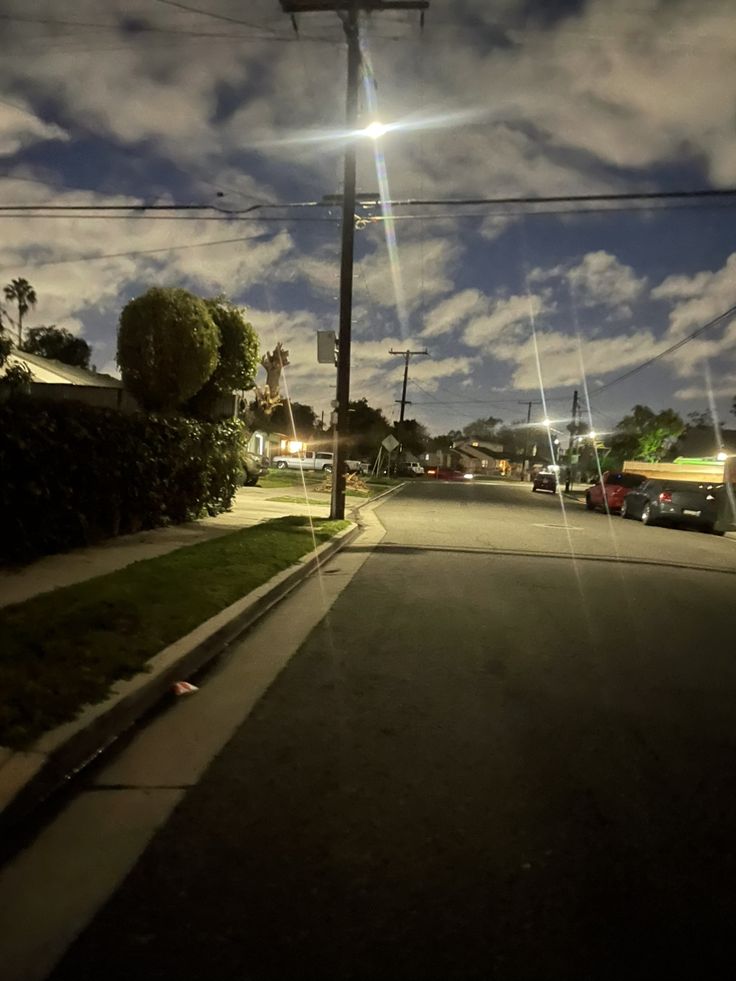 an empty street at night with cars parked on the side and clouds in the sky