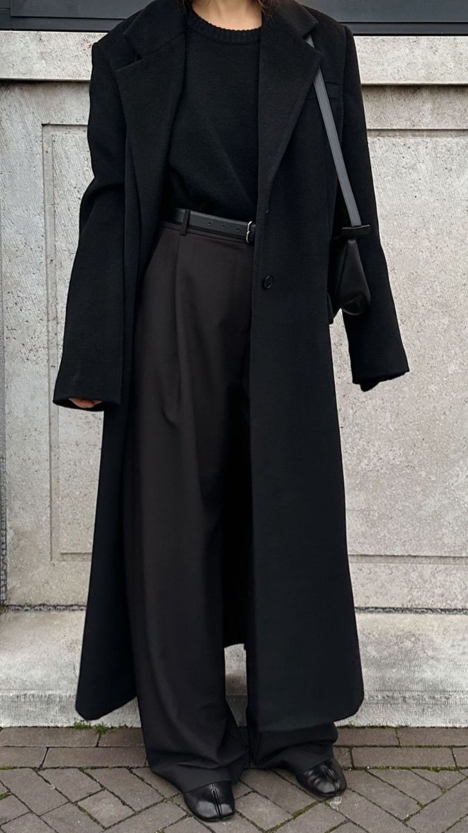 a woman standing in front of a wall wearing a long black coat and wide pants