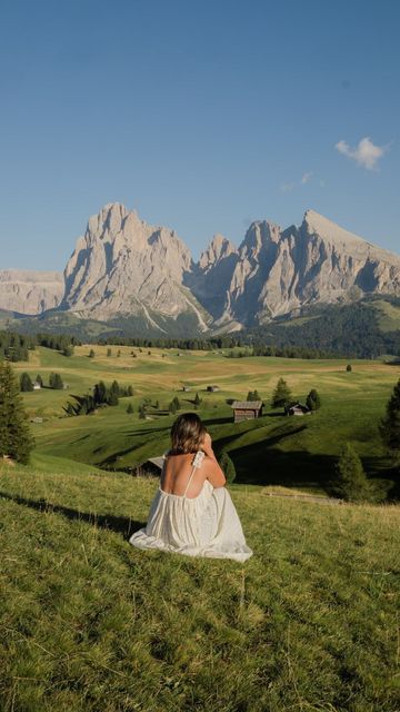 a woman sitting on top of a lush green field next to mountains in the background