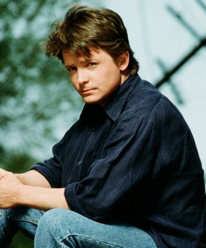 a young man sitting on top of a wooden bench next to a tree and fence