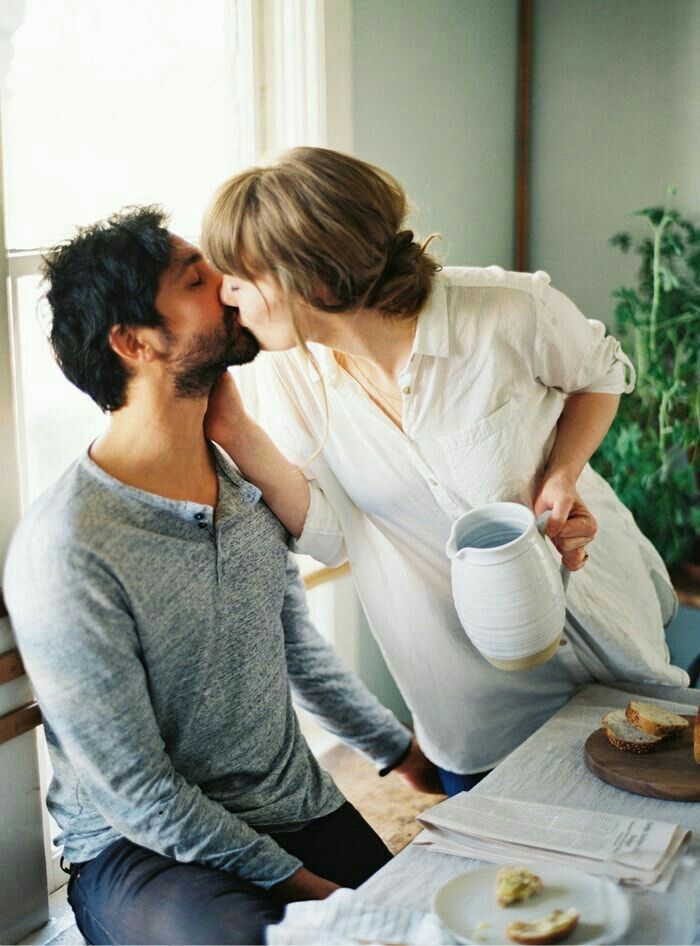 a man and woman kissing each other while sitting at a table in front of a window