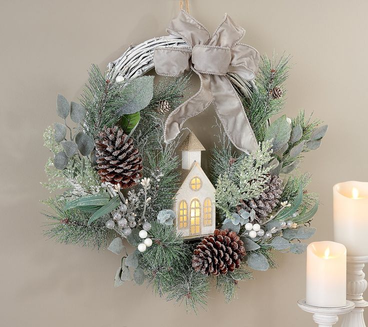 a wreath with pine cones, greenery and a small house on it next to two candles