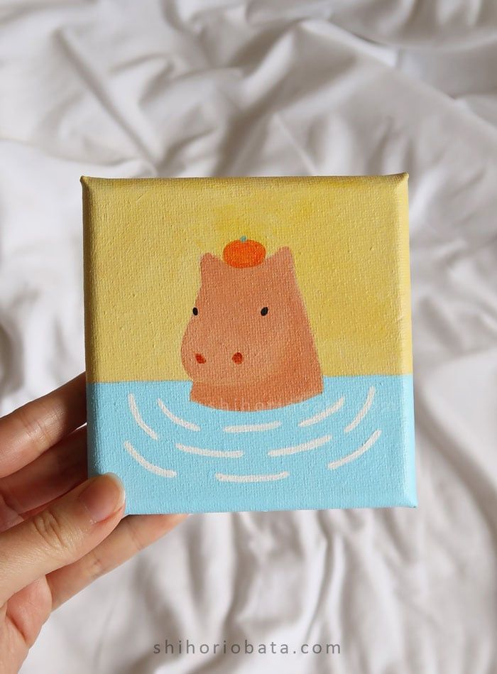 a hand holding up a small painting of a hippo in the water with an orange on its head