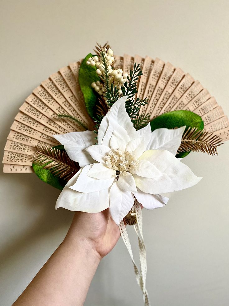 a hand holding a white flower on top of a green leafed fan with greenery