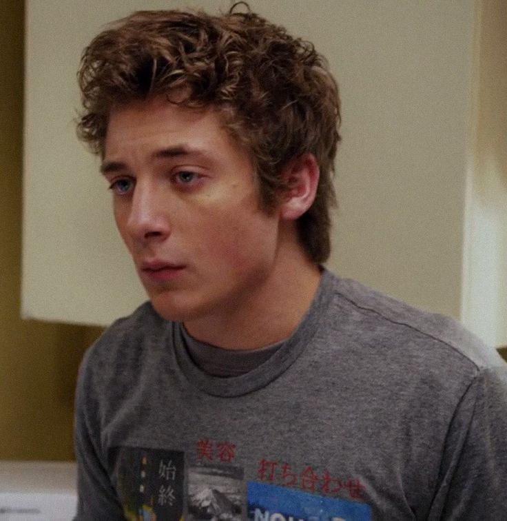 a young man with curly hair wearing a gray shirt and looking at his cell phone