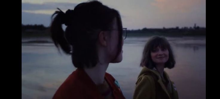 two women standing next to each other near the water at sunset or dawn with clouds in the sky