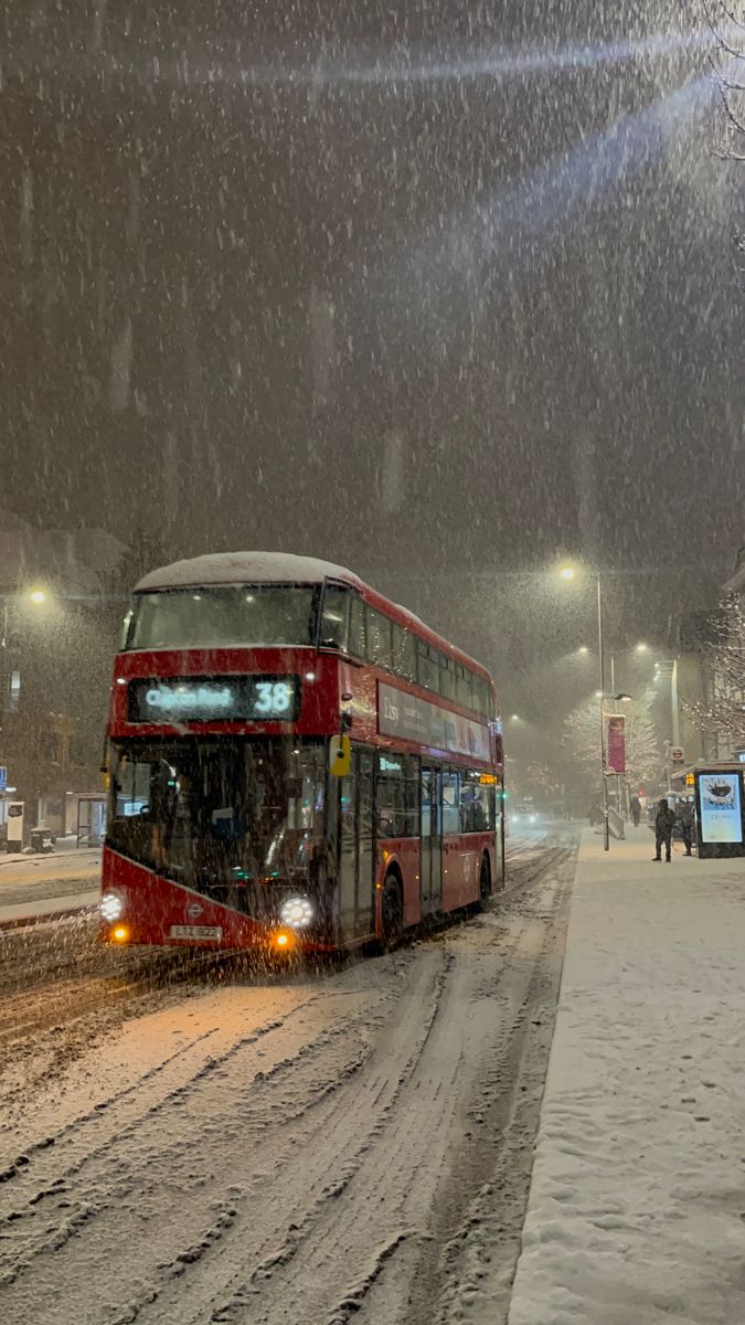 a red double decker bus driving down a snow covered street at night with lights on