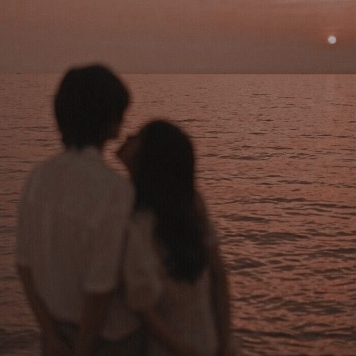 two people standing next to each other looking at the ocean with an orange sunset in the background