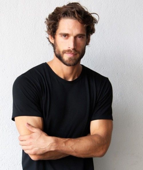 a man with his arms crossed standing in front of a white wall wearing a black t - shirt