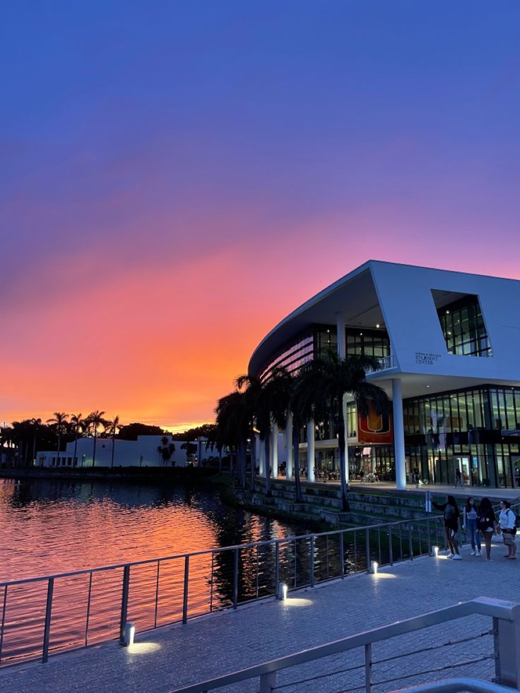 people are walking along the water at sunset near a large building with lights on it