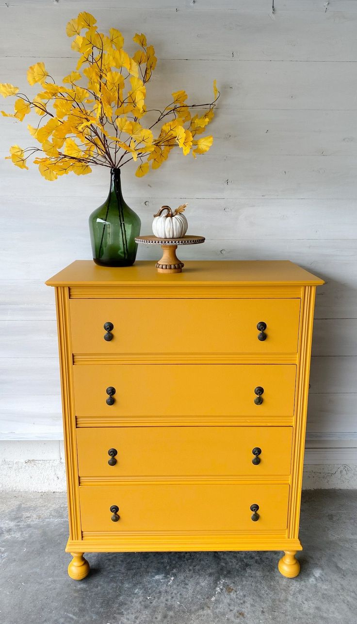 a yellow chest of drawers with flowers and a green vase on top, against a white wall