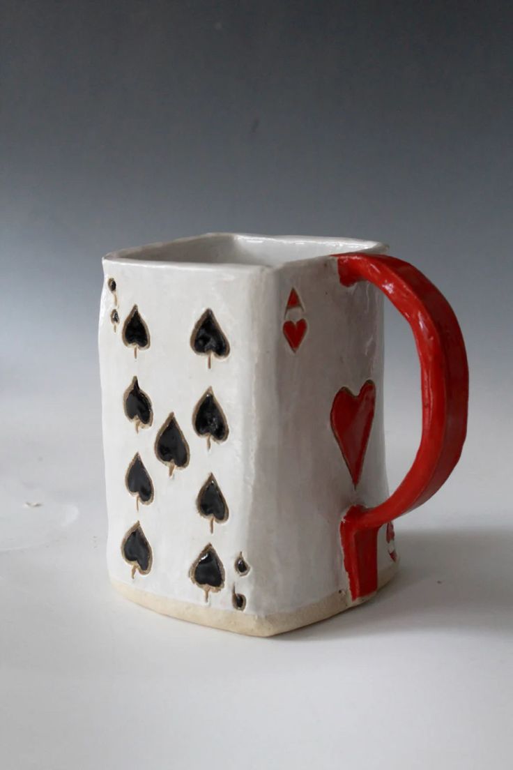 a ceramic mug with playing cards painted on the side and red handles, sitting on a white surface