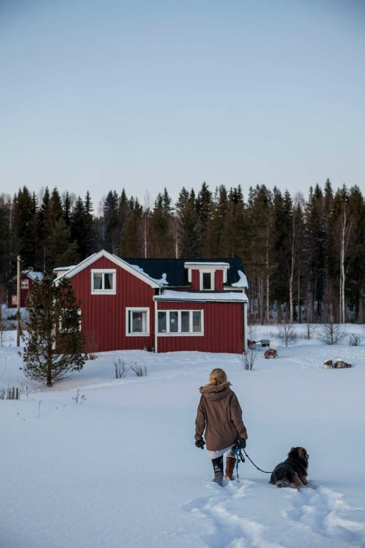 a woman walking her dog through the snow in front of a red house with white windows