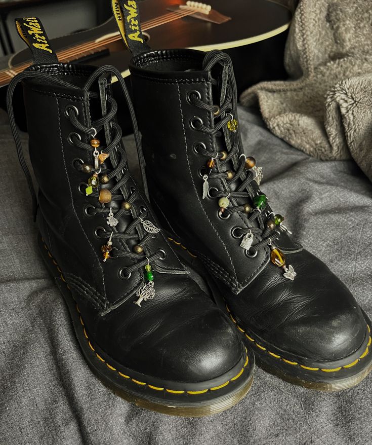 A pair of doc martens with green, brown, and silver charms and beads sitting on grey fabric with a black acoustic guitar in the background Charms On Docs, Outfit W Doc Martens, Diy Doc Martens Charms, Doc Martens With Beads, Decorate Doc Martens, Decorating Doc Martens, Charms On Shoe Laces, Star Shoe Lace Pattern Doc Martens, Doc Martens Beads On Laces