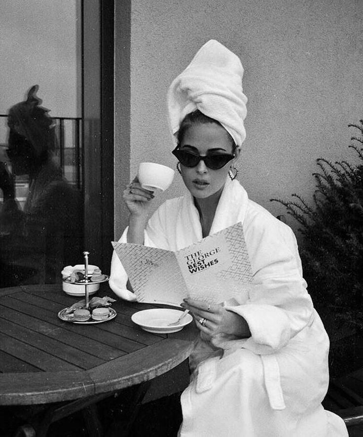 a woman in a bathrobe sitting at a table drinking coffee and reading a book