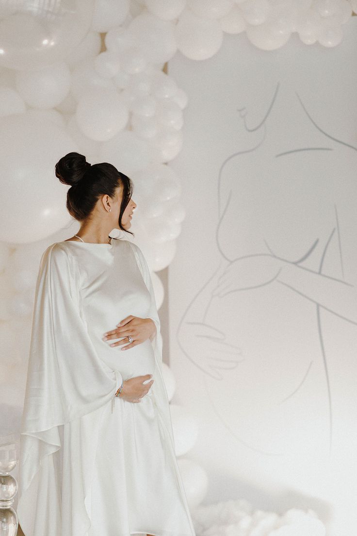 a pregnant woman standing in front of balloons with her belly wrapped around her waist and wearing a white dress