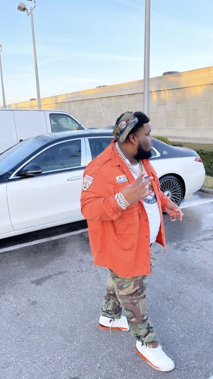 a man in an orange jacket and camo pants standing next to a white car