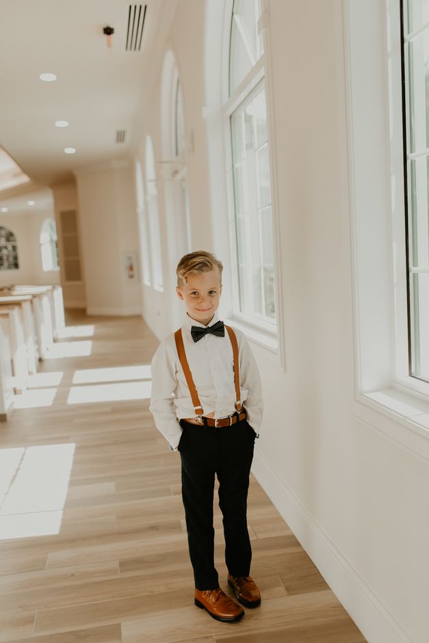 a young boy wearing a bow tie and suspenders standing in front of a window