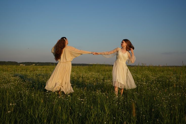 two women in long dresses are holding hands and walking through tall grass on a sunny day