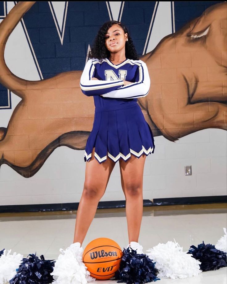 a cheerleader posing with her basketball and pom poms in front of a mural