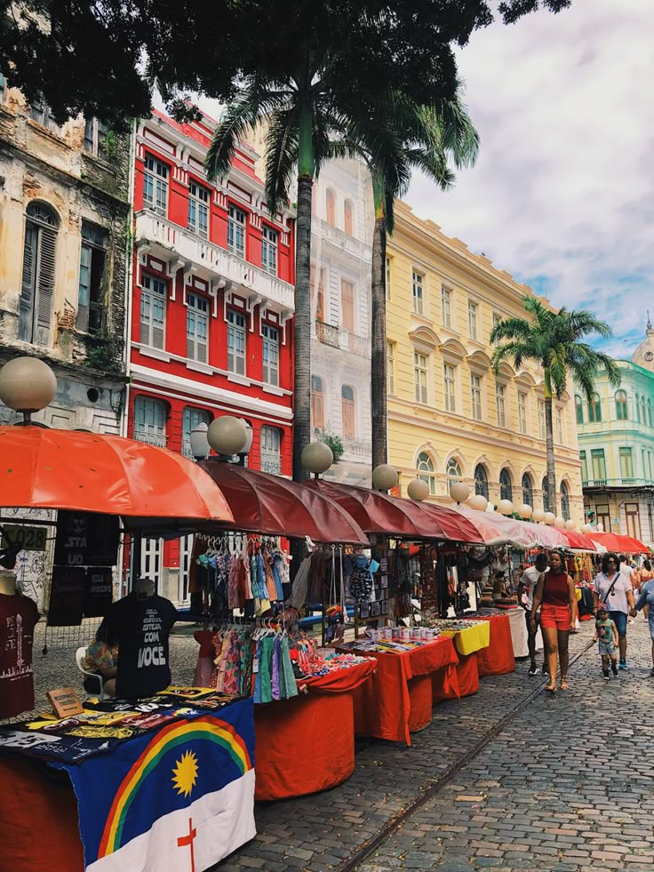 people are walking around an outdoor market with red and yellow umbrellas on the tables