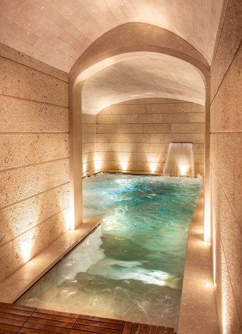 an indoor swimming pool surrounded by stone walls and flooring with lights on either side