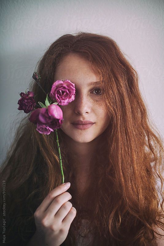 a woman with long red hair holding flowers in front of her face and looking at the camera