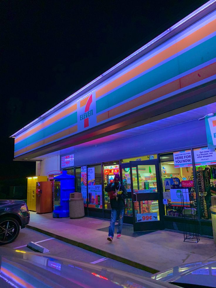 a person standing in front of a store at night with the lights on and cars parked outside