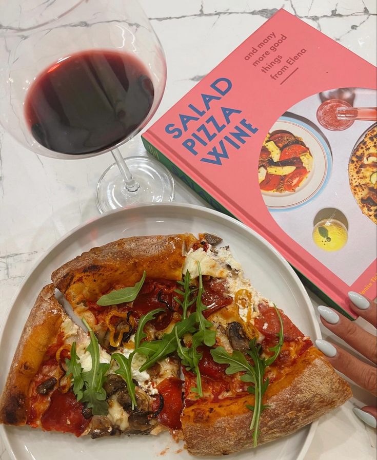 a slice of pizza on a plate next to a glass of red wine and a cookbook