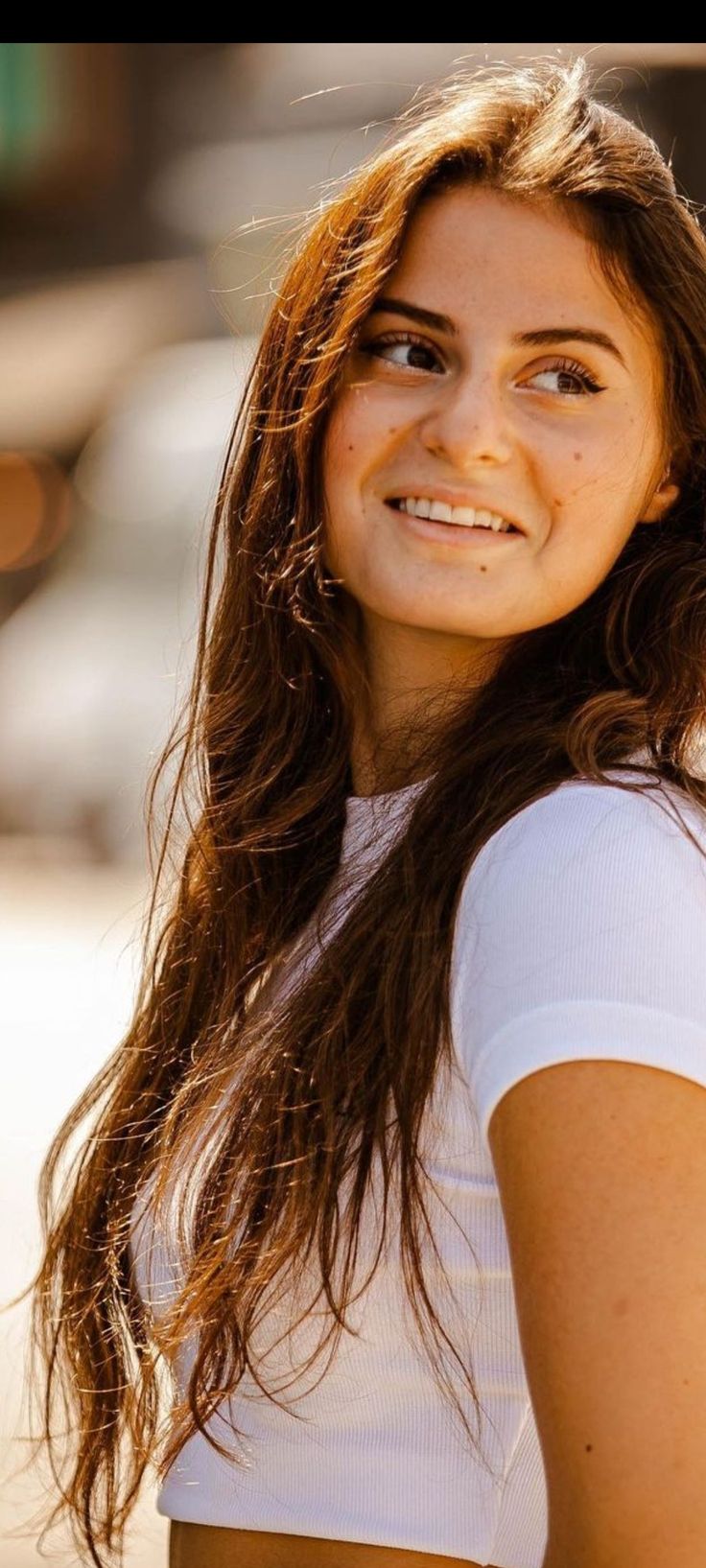 a woman with long brown hair and white shirt smiling at the camera while holding a cell phone in her hand