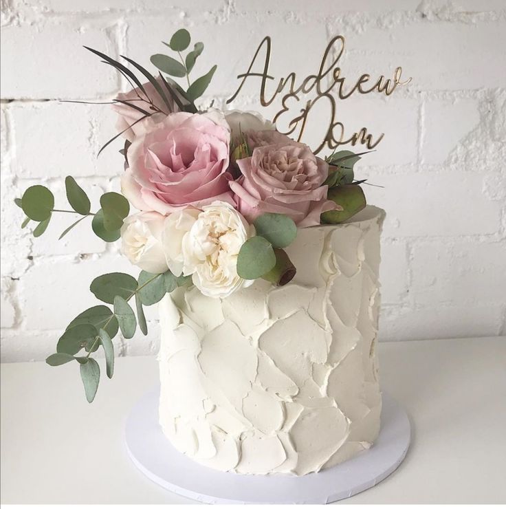 a white cake with pink flowers and greenery on top, sitting on a table in front of a brick wall