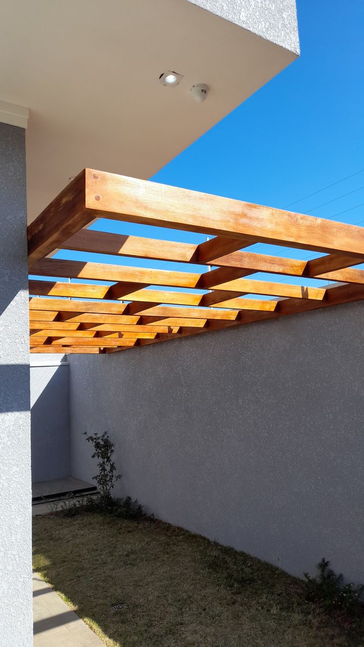 a wooden pergolated roof on the side of a white wall next to a grass area