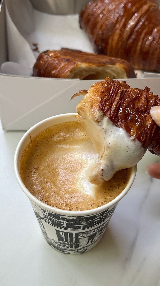 a person is dipping some food into a coffee cup with croissants in the background