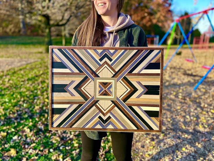 a woman holding up a wooden art piece in front of a playground with trees and grass