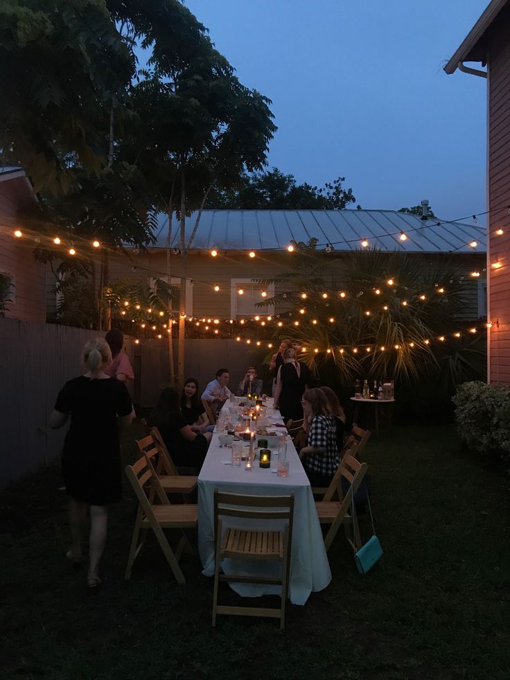 a group of people sitting around a dinner table in the backyard at night with string lights