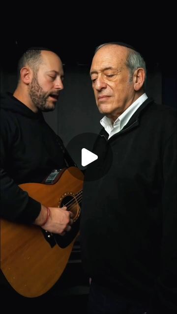 two men standing next to each other while one plays an acoustic guitar and the other sings