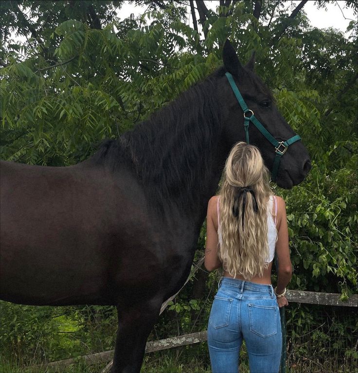 a woman standing next to a black horse in front of some bushes and trees with her back turned towards the camera